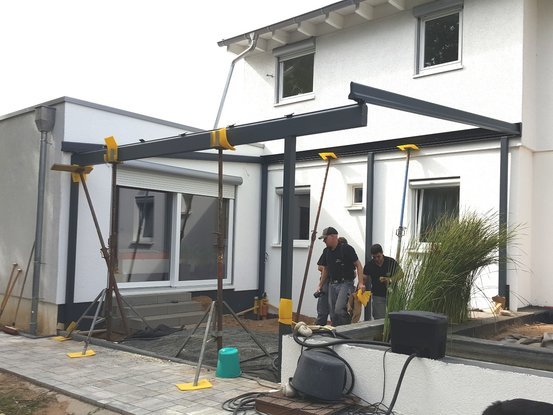 Zwei Handwerker montieren eine Terrassenüberdachung aus Metall an einem modernen Haus.